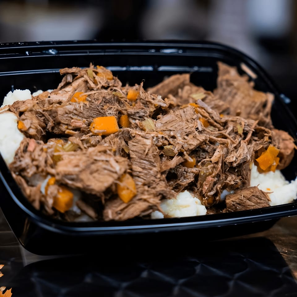 Shredded Italian beef with diced carrots and mashed potatoes in a black takeout container.