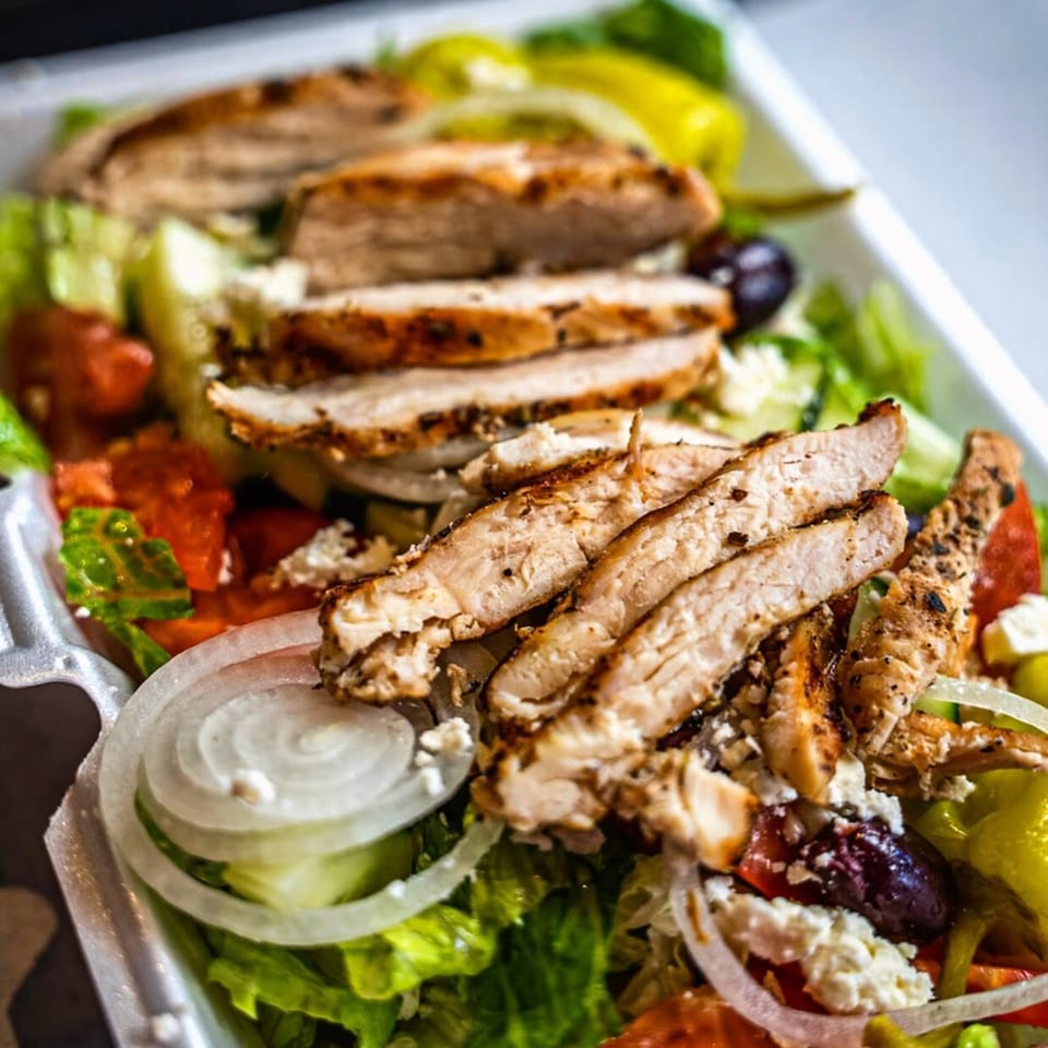 Grilled chicken salad with lettuce, tomatoes, onions, olives, and feta cheese in a white container.